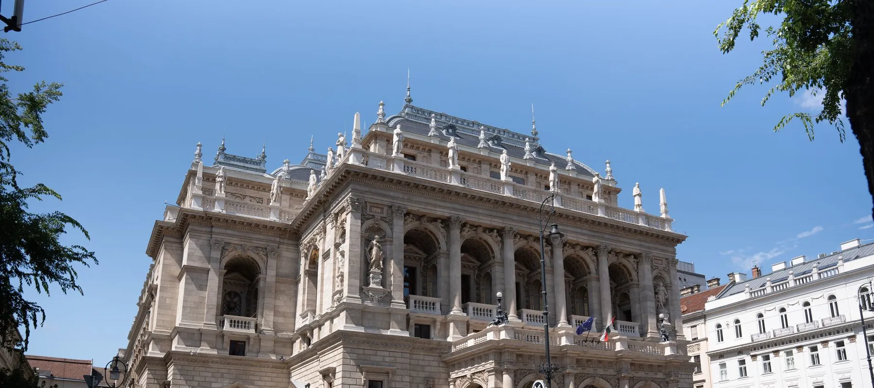 The front of the Hungarian State Opera House on Andrássy út under a blue sky, its roofline lined with statues
