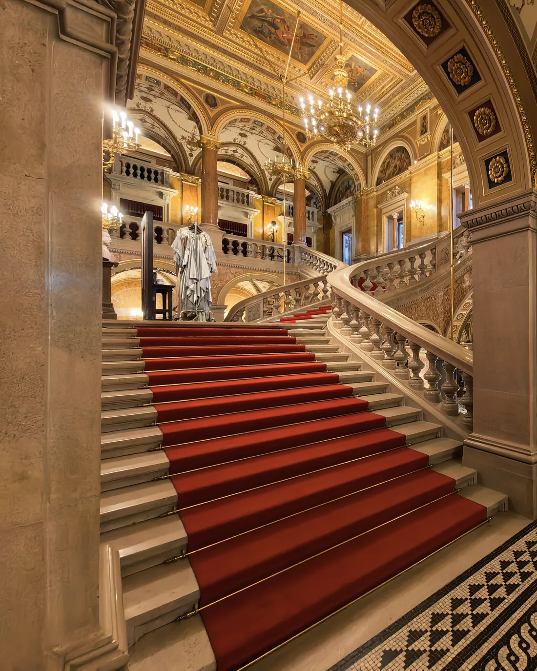 A red-carpeted stone staircase with a carved balustrade rising beneath painted vaults inside the Opera House